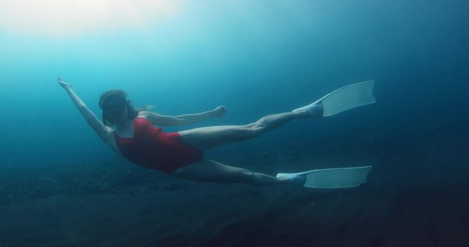 Beauty woman freediver in red bikini with diving fins swims underwater in blue ocean.