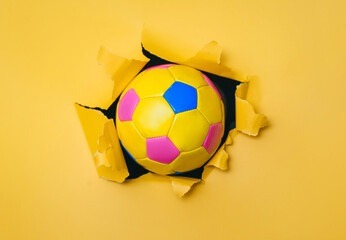 Small colorful children's soccer ball breaks through yellow paper wall. Concept of early age sports, active and mobile team games.