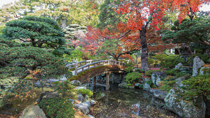 A tranquil scene of a wooden bridge over the Onnai Pond at Kyoto Imperial Palace, surrounded by lush greenery and autumn colors. A peaceful oasis. Kyoto, Japan.