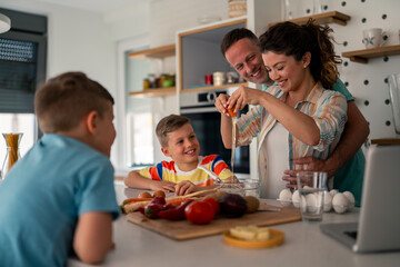 Caucasian family prepares meal in bright kitchen while children learn cooking basics using laptop recipe guide on counter.
