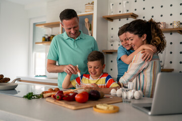 Caucasian parents in casual attire guide their sons through meal preparation activities using kitchen tools and laptop in their modern domestic space.