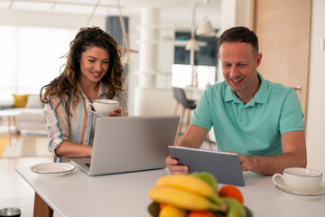 Caucasian couple in casual attire using digital devices while enjoying breakfast at home, surrounded by fresh fruit and coffee cups.