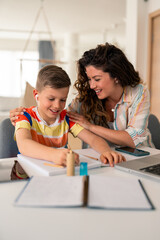 Caucasian mother in striped shirt assists elementary-age son with homework assignment at home study...