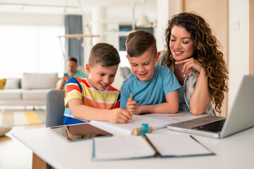 Caucasian mother with curly hair assists two elementary-aged boys with schoolwork using laptop and notebooks in bright home office setting.