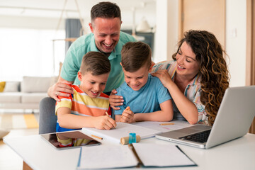 Caucasian parents assist two elementary-aged boys with schoolwork at dining table, using laptop and...