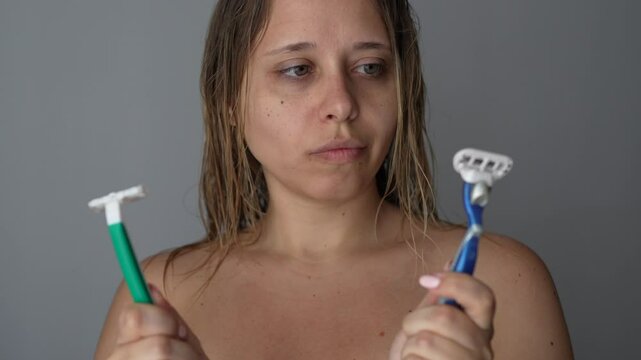 Young caucasian blonde woman holding two types of razors for shaving choosing what to use on dark grey background. Disposable and reusable shavers. Comparison, difference. Getting rid of unwanted hair