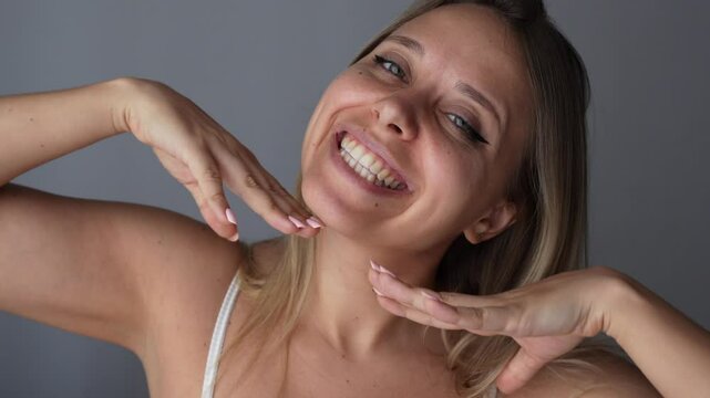 Young caucasian smiling blonde woman showing dimples on her cheek with her hands on a dark grey background. The girl with beautiful smile. Close up