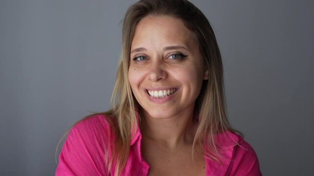 Young cheerful caucasian blonde woman in a pink shirt with dimples on cheeks turns her head and smiles on a dark gray background. Beautiful smile. A lot of moles on the face