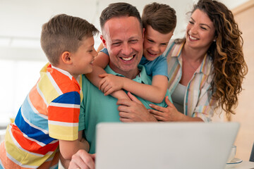 Caucasian professional father in casual attire works on laptop from home while family shares loving moment through playful interaction in bright interior setting.
