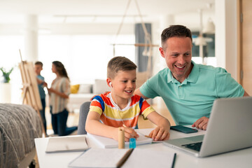 Fototapeta premium Caucasian father in mint polo shirt assists elementary-age son with schoolwork at home, using laptop while women converse in background.