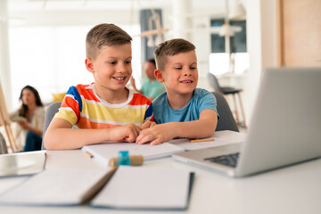 Two Caucasian brothers in casual attire collaborate on schoolwork using laptop and notebooks at bright home study space.