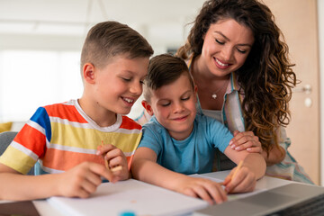 Caucasian mother with curly hair assists two elementary-age boys with homework assignments at dining table in comfortable home environment.