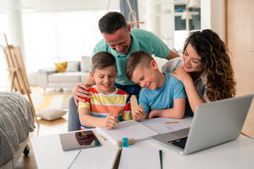 Caucasian parents assist two young boys with educational activities at home dining table, using laptop and writing materials in bright modern interior setting.