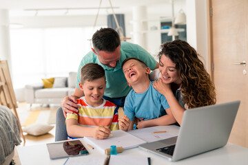 Caucasian parents assist two young boys with schoolwork at dining table, using laptop and writing materials in bright home setting, sharing joyful learning moment.