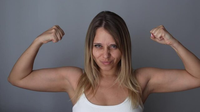Young cheerful caucasian strong fit blonde woman raising arms and showing biceps on a dark grey background. Feminism, girl power, equal women's rights, independence, sports concept