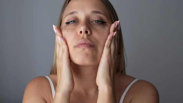 Young blonde woman massaging her face quickly slapping herself on the cheeks to improve blood circulation, relax the muscles, tighten skin on a dark grey background. Cosmetology and beauty. Facelift