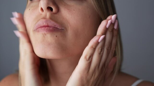 Close-up of young blonde woman massaging the lower part of her face slapping herself on the cheeks on a dark grey background. Rejuvenation, facelift, facefitness, skin care. Cosmetology and beauty