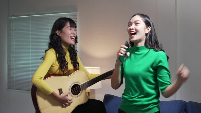 Two cheerful Asian women playing acoustic guitar and singing together, sharing musical connection in cozy living room setting