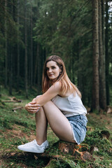 young woman sitting on a stump in the forest. She is wearing a white T-shirt and denim shorts. She has white sneakers on her feet.