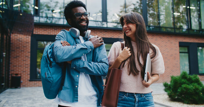 Two multi-ethnic teachers walking away from school. Telling each other stories about past working day. Smiling while talking. Modern building in blurred background. Happy about finishing job early.