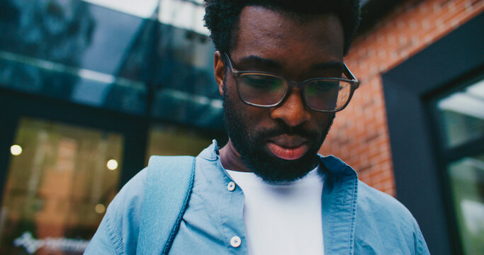 Bearded African American student with backpack standing in front of brick wall. Smiling to himself. Browsing notifications on screen. Opening messages in social media. Wearing headphones on neck.