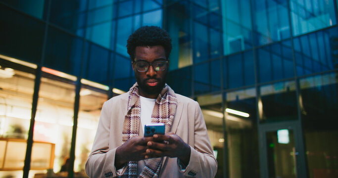 Camera focus on handsome African American male with glasses slowly walking away from business center. Looking at smartphone. Reading news or message in social media. Cold weather. Evening.