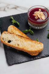 Delicious beetroot cream soup is served with almond flakes, toasted slices of bread and basil leaves on black slate plate on white marble table. Restaurant or cafe menu, cookbook recipe