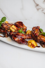 Glazed chicken wings are served on a white rectangular plate, garnished with mango chunks, sesame seeds, fresh basil leaves, and a drizzle of sauce. Restaurant or cafe menu, cookbook recipe