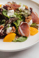 Fresh salad with parma ham, melon slices, walnuts, goat cheese and mixed greens, served on a white plate on a marble table. Restaurant or cafe menu, cookbook recipe