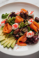 Elegant smoked salmon salad served with fresh avocado slices, mixed greens, olives, cream cheese, and vibrant red fish roe, presented on a white plate. Restaurant or cafe menu, cookbook recipe