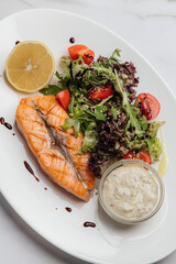 Grilled salmon fillet with fresh salad, lemon slice, tartar sauce, and balsamic glaze drops, placed on a marble table, creating a delicious and healthy meal. Restaurant or cafe menu, cookbook recipe
