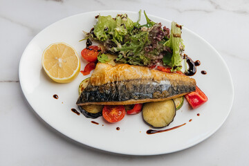 Grilled mackerel fillet is served on a white oval plate with grilled vegetables, fresh salad, lemon slice, and balsamic sauce on a marble table. Restaurant or cafe menu, cookbook recipe