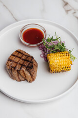 Grilled beef steak resting on a white plate, accompanied by vibrant grilled corn, sliced red onion, fresh microgreens, and a drizzle of savory sauce. Restaurant or cafe menu, cookbook recipe