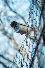 Obraz premium A surveillance camera is mounted on a pole, overlooking a fenced area. Barbed wire coils around the fence, adding a layer of security. Bright sky creates a stark contrast with the scene
