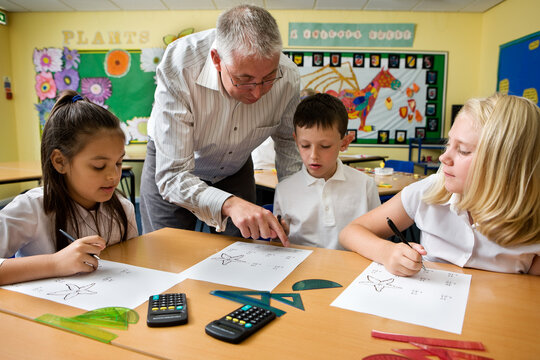 Junior School: Maths Advice. A group of young primary school children working at their desk in class on a maths project while teacher offers some help.