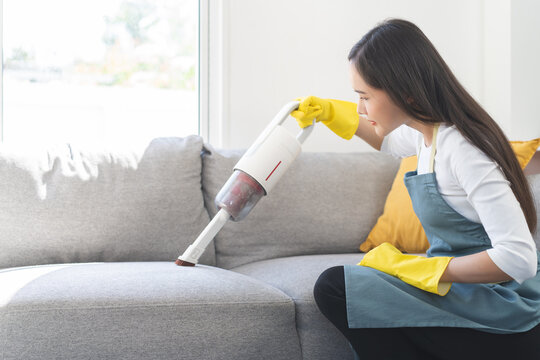Household cleaning concept, asian young woman hand of woman vacuuming dust and dirt on sofa, doing housework and chores cleanup furniture in living room at home. House cleaning with handheld hoover.