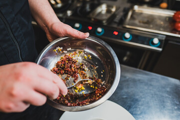 A chef mixes colorful ingredients in a stainless steel bowl, showcasing culinary preparation in a professional kitchen, with blurred appliances in the background.