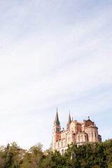 A view from Covadonga in Spain. Looking up at an ancient cathedral rising from the green trees and glowing in the afternoon sunlight beneath a blue sky with thin white clouds.