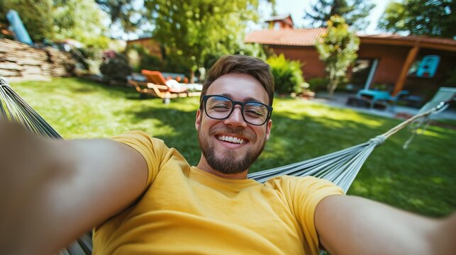 A smiling man relaxes in a hammock, surrounded by a lush green garden on a bright sunny day. His carefree expression reflects joy as he captures the moment with a selfie
