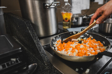 A hand stirs diced onions and carrots in a skillet on a stovetop, with blurred pots and cooking oil in the background, showcasing a dynamic kitchen setting.