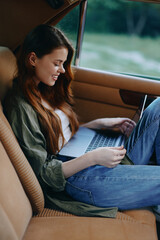 Young woman with long red hair sitting in a vintage car, smiling while working on a laptop, wearing a casual olive green jacket and jeans.