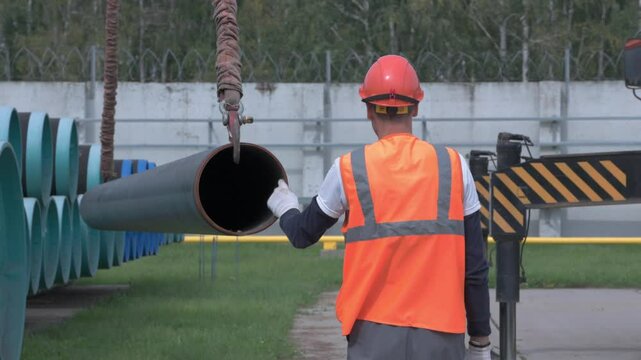Construction worker guides heavy pipe during crane lifting on industrial site.