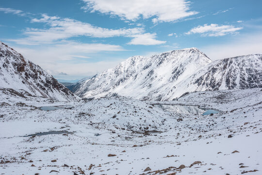 Cold snow-white landscape of snowy valley with alpine lakes against snow-covered rocky mountain range under clouds in blue sky. Glacial lakes, pure white snow and stones in high mountains in sunny day