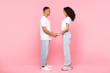 Black man and woman holding hands and looking at each other, standing on pink background, full length, side view