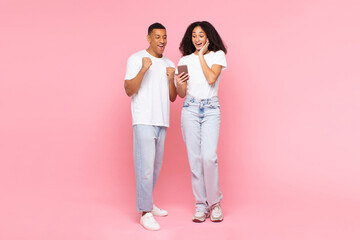 Joyful Latin couple holding cellphone, reading great news, shaking fists in joy over pink studio background, full length shot