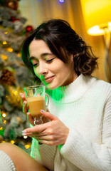 Woman with drink by tree. A woman in a cozy sweater sips a warm beverage while sitting next to a beautifully decorated holiday tree.