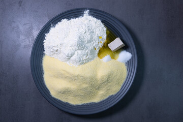 Ingredients for homemade bread or dough: flour, cornmeal, yeast, salt, sugar, and olive oil arranged on a dark plate.
