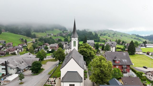 Drone flight above the village centre of Wildhaus, a charming village near the Alpstein mountain range. Small town centre with church and houses. Surrounded with beautiful Swiss mountains. Cloudy summ