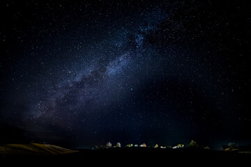 Naklejka premium Stunning panorama of the starry night sky with the Milky Way over an illuminated camp in the middle of the Sahara desert in Morocco. Adventure and freedom travel concept
