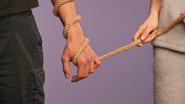 Man hand tied with rope, woman holding the rope as leash, abusive dominated feministic relationships concept, isolated on purple studio background, close up shot.